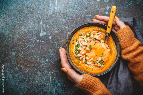 Female hands in yellow knitted sweater holding a bowl with pumpkin cream soup on dark stone background with spoon decorated with peanuts, top view. Autumn cozy dinner concept 
