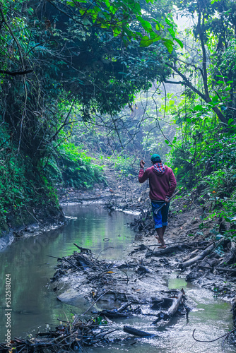 Man walking by the creek in the forest with their shoes in hand. Hikers hiking in the forest. Hiker walking in the forest beside the mountain river. © NadimMahmudHimu