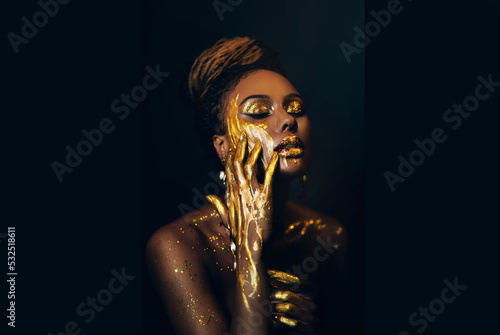 Artwork portrait closeup fantasy african woman face in gold paint. Golden shiny skin. Fashion model girl goddess. hand fingers in metallic liquid drop. Arab royal style professional glamorous makeup