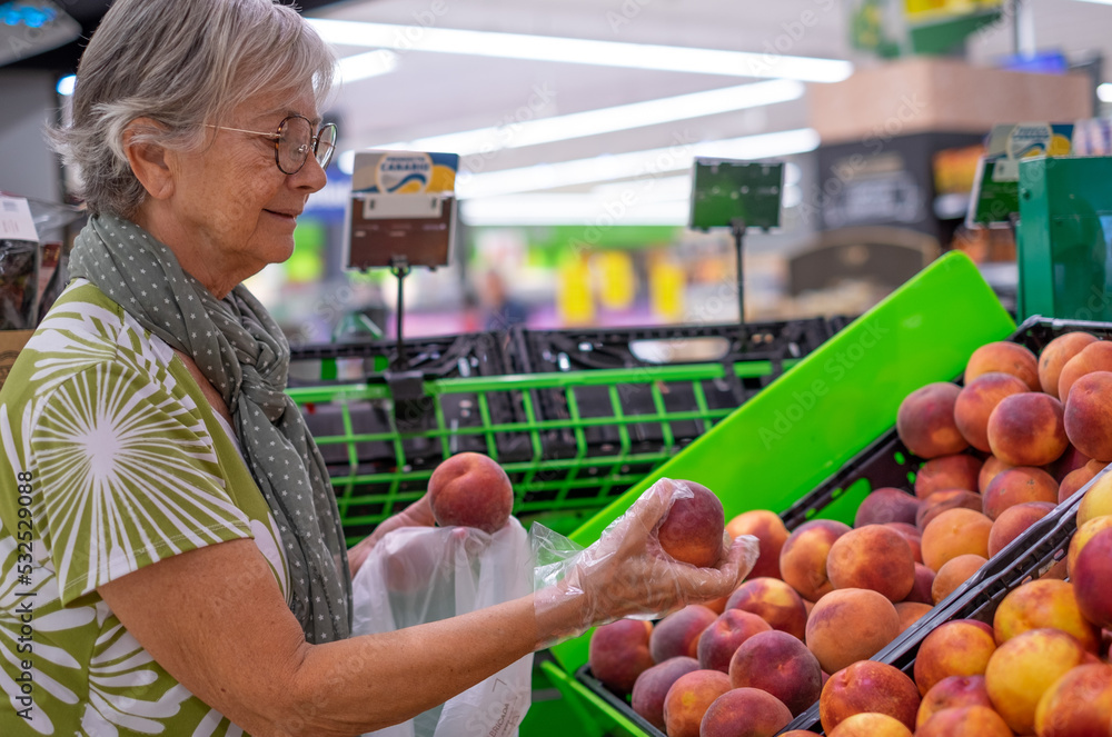 Obraz premium Senior woman wearing protective gloves selecting fresh peaches in the supermarket - consumerism concept, rising prices, inflation