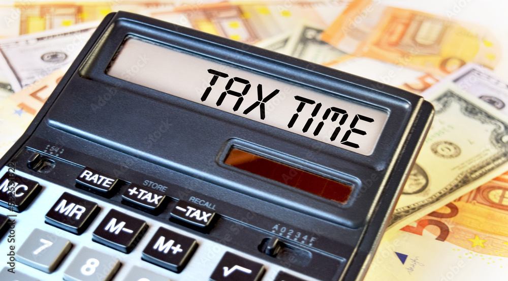 TAX TIME text written on a calculator next to the background of euro banknotes. Finance or business concept. TAX TIME text written on a calculator next to the background of euro banknotes. Finance or business concept.
