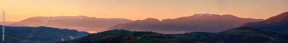 panorama of mountainous countryside at dawn. hills with trees and rural fields rolling down into the foggy valley. distant ridge with snow capped tops beneath a sky with clouds in red morning light panorama of mountainous countryside at dawn. hills with trees and rural fields rolling down into the foggy valley. distant ridge with snow capped tops beneath a sky with clouds in red morning light