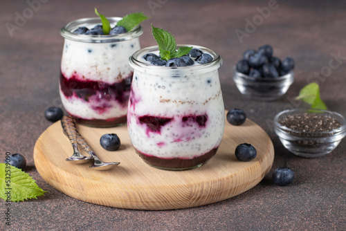 Chia pudding with blueberry and jam in two glass jars on wooden board on brown background