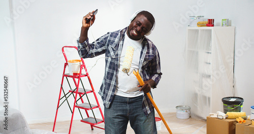 Happy African American handsome young guy dancing with paint brush roller in hand listening to music in headphones enjoying song from smartphone during room reconstruction, home renovation concept