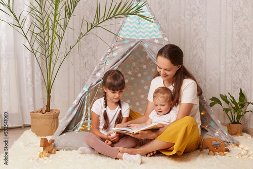 Fotografie Horizontal shot of happy mother wearing white shirt and yellow trousers sitting on floor with her kids and reading book, being interested in interesting plot of fairy tale