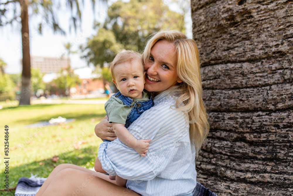 Young Mother Leans Against A Tree In The Park While Holding Her Son Young Mother Leans Against A Tree In The Park While Holding Her Son
