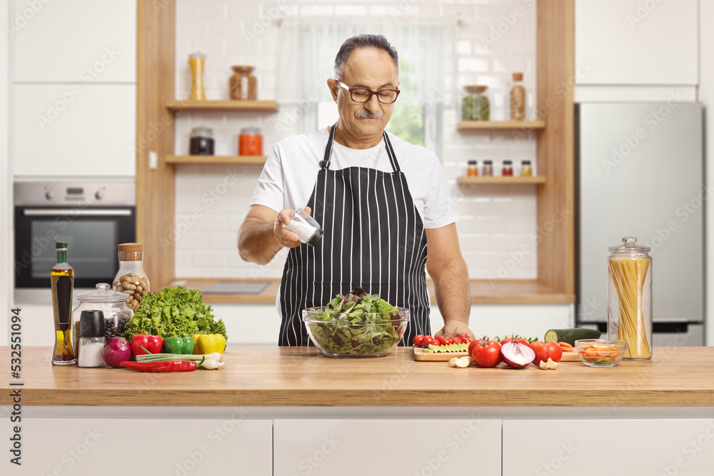Mature man with an apron seasoning a salad with salt on a kitchen counter Mature man with an apron seasoning a salad with salt on a kitchen counter