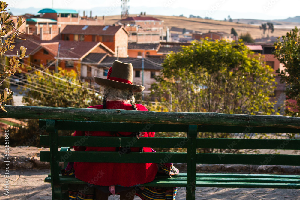 Obraz premium Old woman, in typical Peruvian dress, sitting with her back on a bench in a park with a view of the city of Chinchero, Peru. 