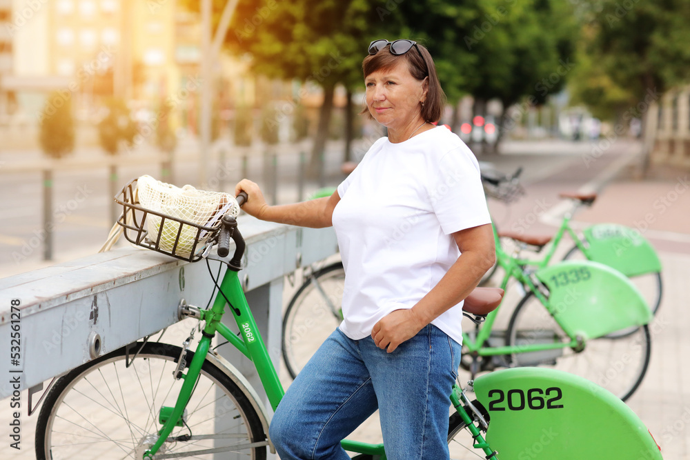 Obraz premium Beautiful mature woman smiling while standing on the street near bicycles. Female in her hand using bike rental service on summer sunny day. Active lifestyle
