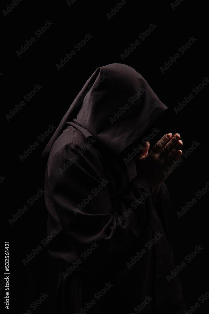 side view of mysterious monk in hood praying isolated on black.