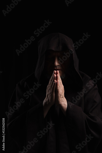 front view of monk in dark hooded cassock praying isolated on black.