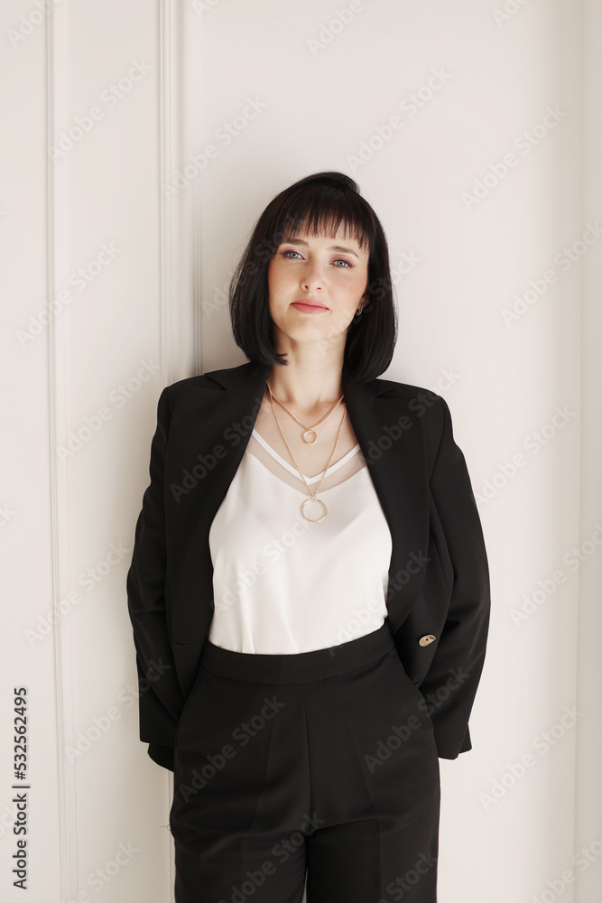 Confident young brunette business woman looking at camera on white wall background. Studio portrait of successful friendly female with short hair in black suit and white blouse with jacket on shoulder