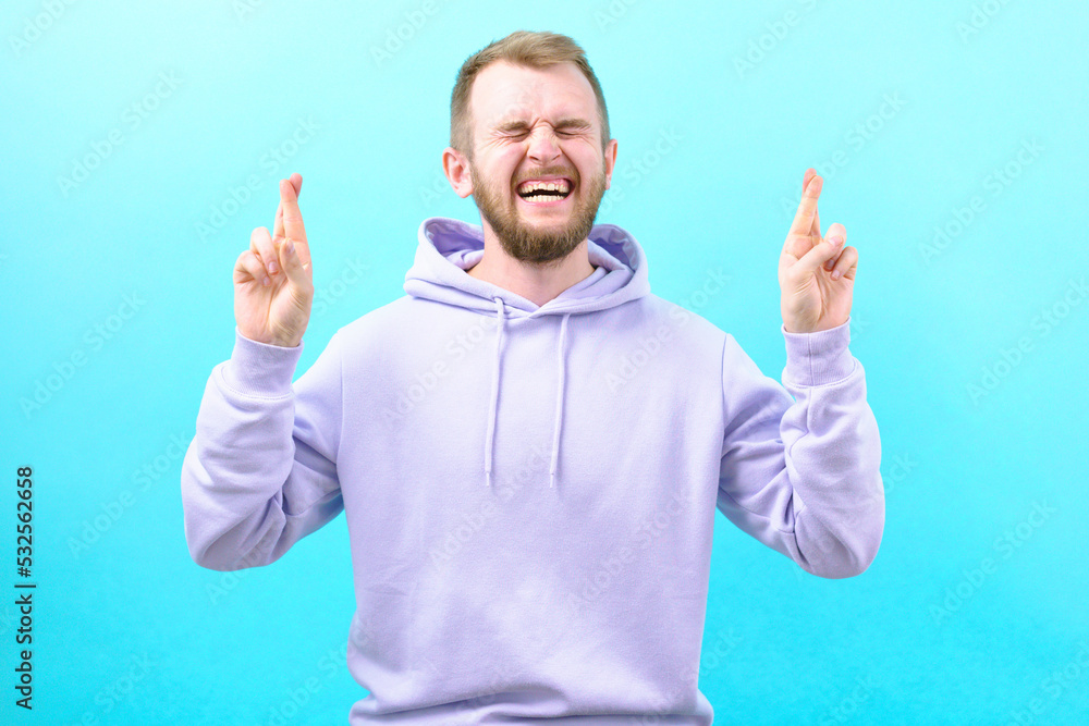 Man with a wry face crossing his fingers on a blue background. Hoping for luck. Panic. Fright. Suffer. Scared. Scary. Feared. Anxious. Frightened. Afraid. Frowning. Stress. Upset. Frustration Man with a wry face crossing his fingers on a blue background. Hoping for luck. Panic. Fright. Suffer. Scared. Scary. Feared. Anxious. Frightened. Afraid. Frowning. Stress. Upset. Frustration
