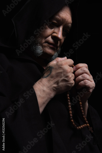 senior tattooed monk in hood holding rosary while praying isolated on black.
