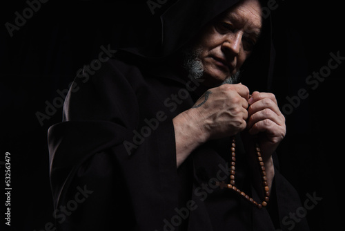 low angle view of monk in hooded robe praying with rosary isolated on black.