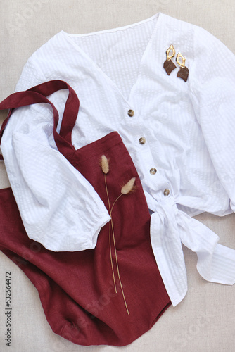 Flat lay of female clothes and accessories - white shirt, burgundy fabric tote bag and oversized earrings. View from above.