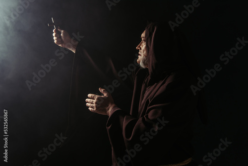 monk in black hooded robe holding holy crucifix in outstretched hand on dark background.