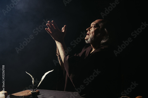 side view of medieval monk praying near inkpot with quills on black background.