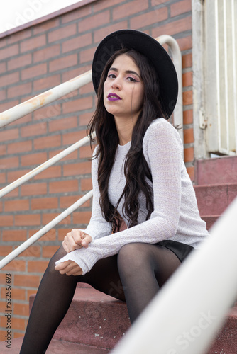 sitting resting on some bleachers, young latin woman wearing casual clothes with hat, lifestyle and beauty, portrait of one person