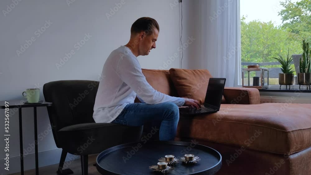 White Caucasian man in casual clothes is working for his business from home in the living room with his laptop and is thoughtfully looking up and is showing a happy face. There are no trademarks in th White Caucasian man in casual clothes is working for his business from home in the living room with his laptop and is thoughtfully looking up and is showing a happy face. There are no trademarks in th