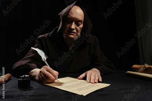 priest in hooded robe writing chronicle on parchment isolated on black.