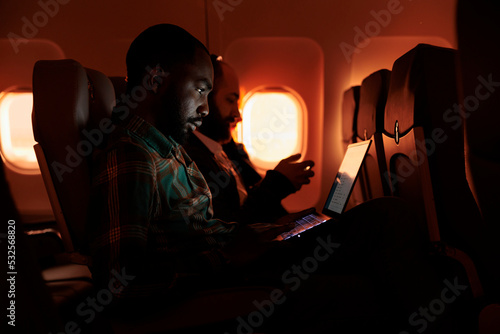Freelancer passenger working on laptop during sunset, flying by airplane on commercial flight with international airline service. Male tourist travelling on work trip or holiday.
