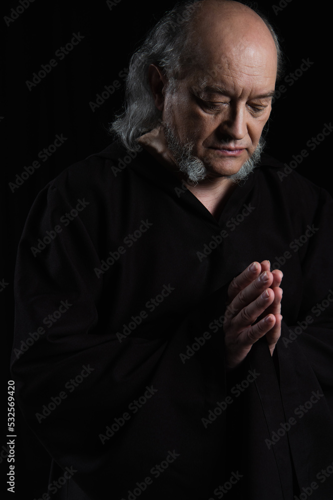 senior medieval monk in dark robe praying isolated on black.