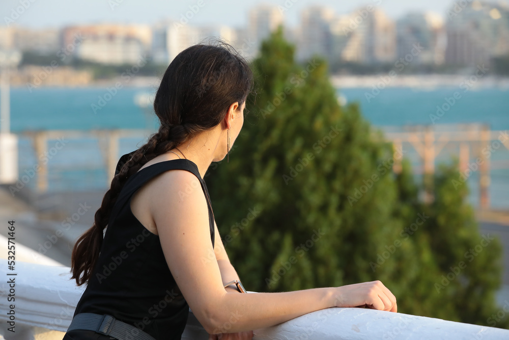 A woman standing on the embankment looks at the sea from the shore.Portrait of a young brunette with a pigtail enjoying the view of nature in a resort town on a summer day