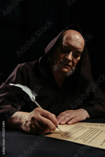 senior medieval priest writing ancient chronicle on parchment isolated on black.