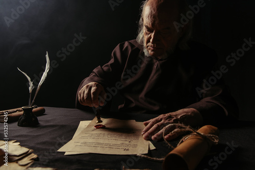 ancient monk stamping manuscript with wax seal near rolled parchment isolated on black.