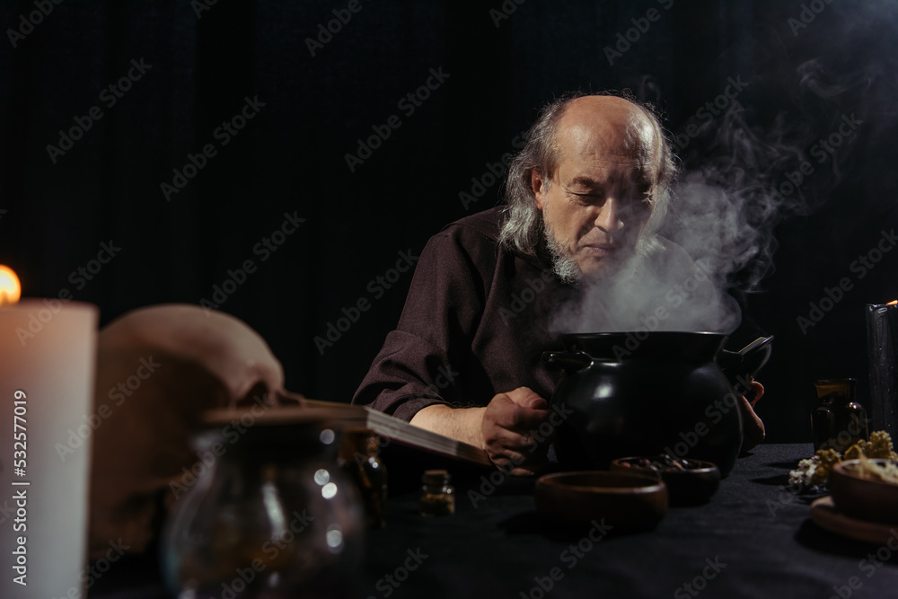 senior alchemist near boiling and steaming pot preparing potion at night isolated on black. senior alchemist near boiling and steaming pot preparing potion at night isolated on black.