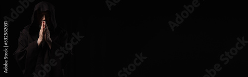 front view of medieval monk in dark hood praying isolated on black, banner.