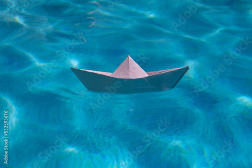 Paper boat sailing on water causing waves and ripples. Paper boat into water. Concept of tourism, travel dreams vacation holiday. Blue water background. © Volodymyr