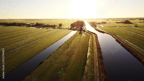 Aerial drone footage of vast green farmland fields in the Netherlands, separated by water canals with a windmill in the middle. Autumn sunrise.