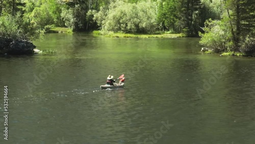 Aerial drone view following tourist with dog in kayak, paddling towards green mountain forest coast on sunny summer day in Sierra Nevada Mountains, California. Traveler on fun weekend water activity