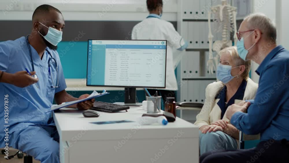Medical worker consulting old woman with physical disability, doing checkup examination at clinic. Nurse talking to paralyzed patient and husband with face masks at appointment. Medical worker consulting old woman with physical disability, doing checkup examination at clinic. Nurse talking to paralyzed patient and husband with face masks at appointment.