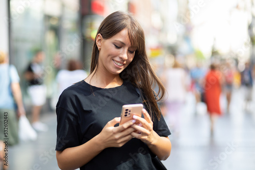 Beautiful smiling woman in black shirt texting on smartphone on street in city center
