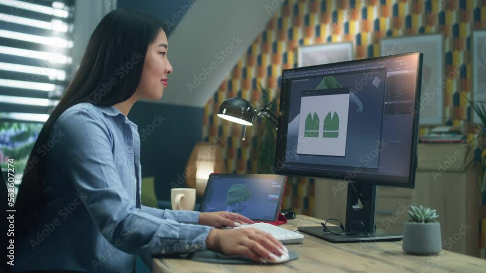Asian woman creating 3D models of clothes on computer and tablet in 3D modeling program, while sitting at the desk and working remotely at home office. Asian woman creating 3D models of clothes on computer and tablet in 3D modeling program, while sitting at the desk and working remotely at home office.