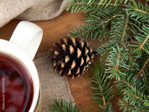 Part of the mug is visible on a wooden table, standing on a linen napkin next to spruce branches and a cone, in the background a linen tablecloth