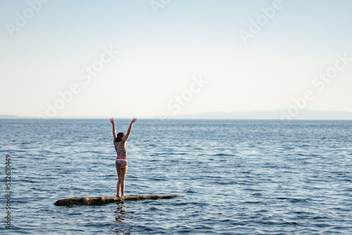 Girl stands on driftwood log in ocean arms raised in victory