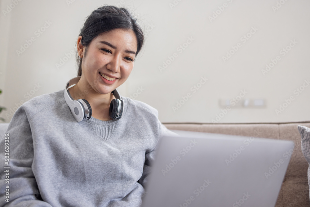 An business Asian woman working at home, chatting online with a colleague on her laptop and wear headphones while on the couch in living room at home. An business Asian woman working at home, chatting online with a colleague on her laptop and wear headphones while on the couch in living room at home.