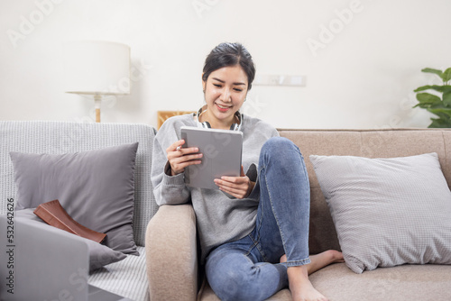 Beautiful Asian woman using digital tablet while headphones sitting on sofa at home