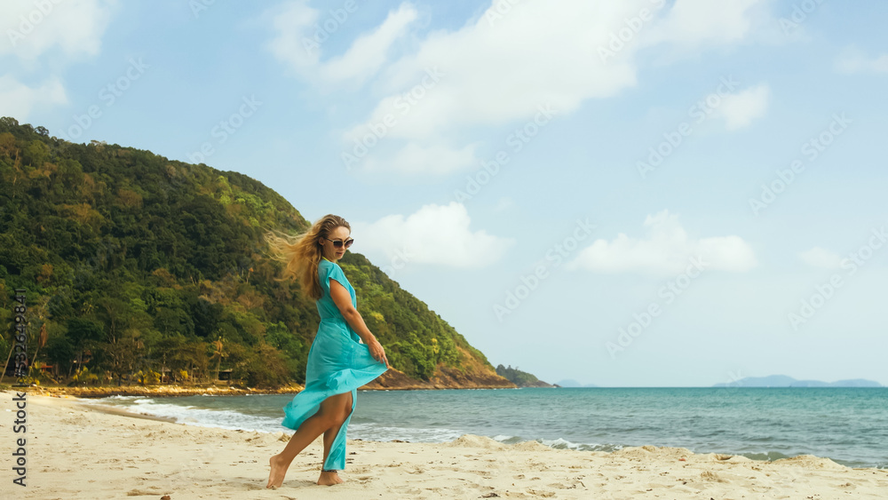 Beautiful barefoot young lady in sunglasses walking on the sunny beach blue silk dress fluttering in the wind great day for walk. Concept rest tropical resort traveling tourism happy summer holidays