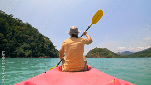 Young man with sunglasses and hat rows pink plastic canoe along sea against green hilly islands with wild jungles. Traveling to tropical countries. Strong guy is sailing on kayak in ocean, back view. © ivandanru