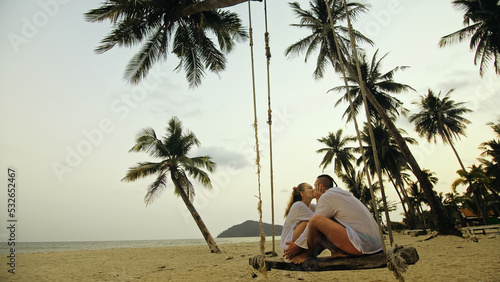 Cute loving couple on the swing on tropical beach. Woman and man in white shirt enjoy gold sunset and life in beaches, palm tree. Evening warm sunset. Summer holiday vacation tropical tourism concept