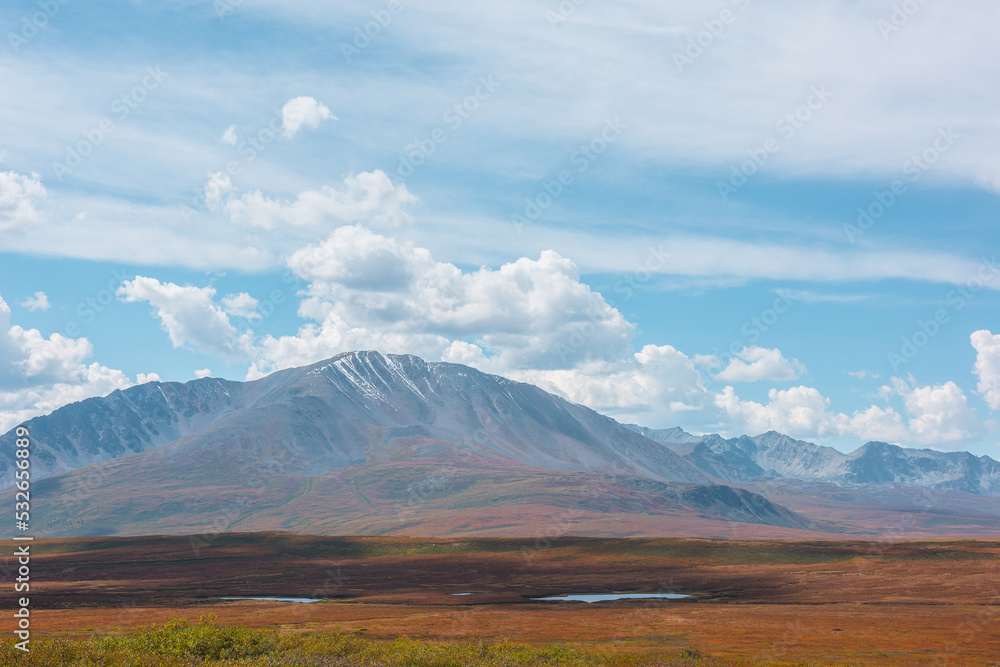Fototapeta premium Motley autumn landscape with sunlit high mountain plateau with lakes and beautiful mountain top under dramatic cloudy sky. Vivid autumn colors in mountains. Shadows of clouds in changeable weather.