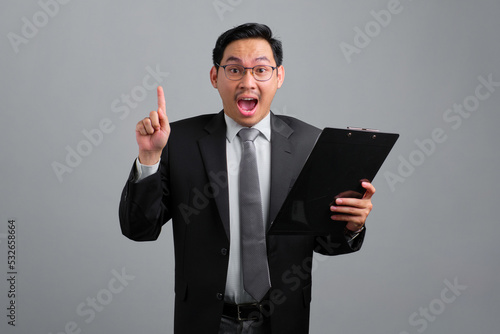Portrait of excited handsome young businessman in formal suit with a clipboard having idea and pointing upwards isolated on grey background