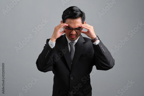 Frustrated handsome young businessman in formal suit suffering from headache because migraine isolated on grey background
