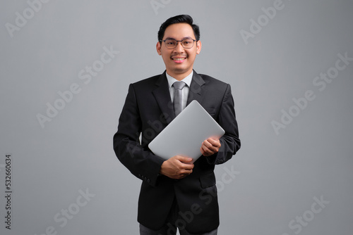 Portrait of smiling handsome young businessman in formal suit and glasses holding closed laptop isolated on grey background