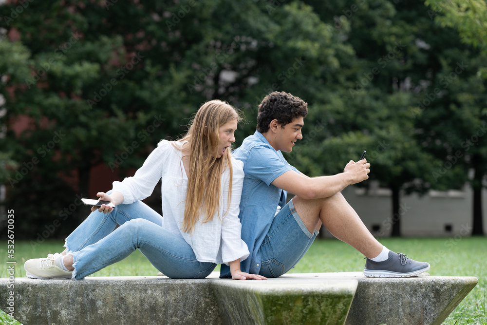 Couple sitting back to back outdoors in a park. Cheating concept. Girl spying phone to girlfriend.